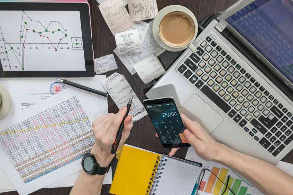 IT costs - hands holding phone calculator on messy desk with charts, papers, and laptop